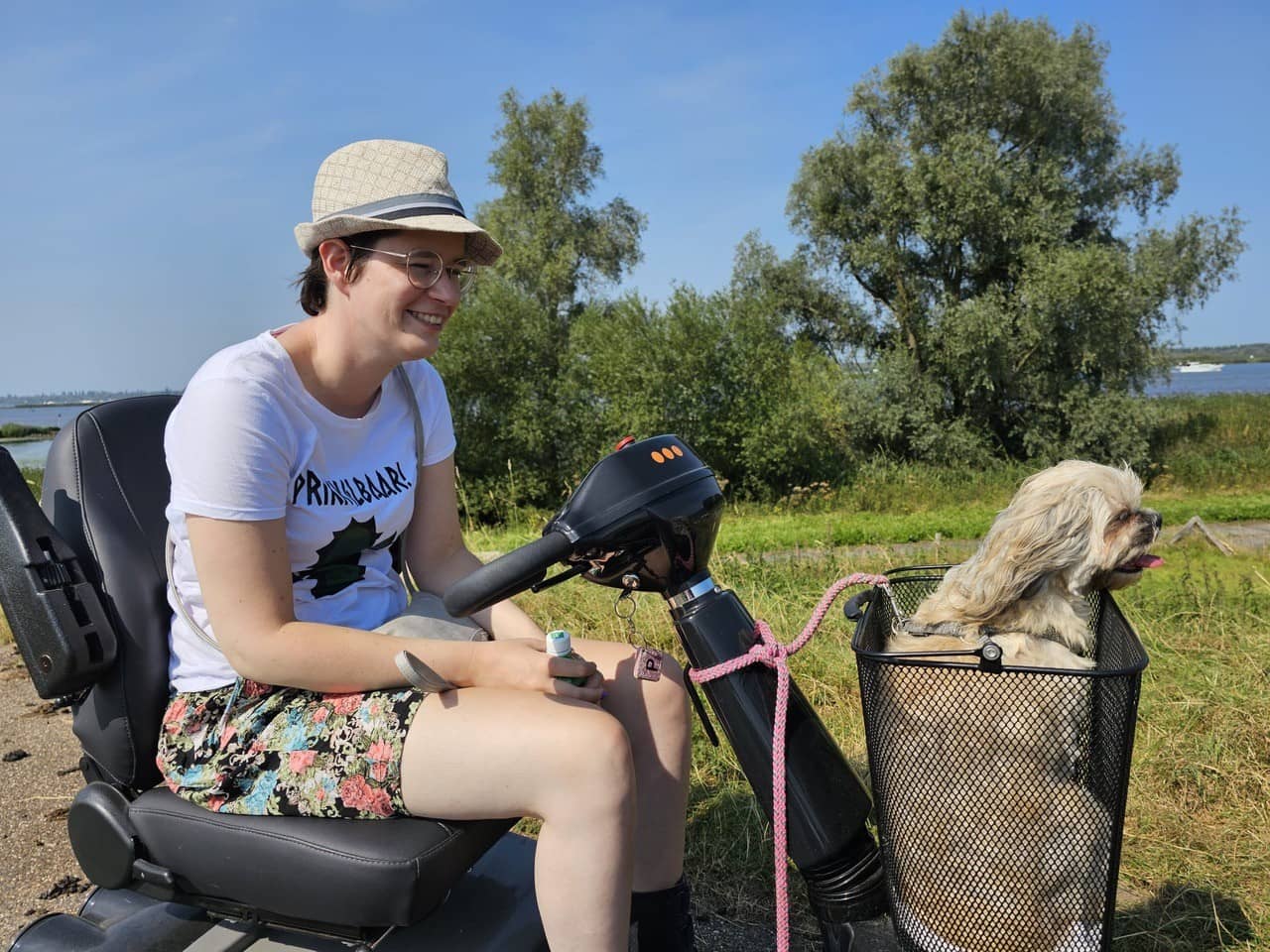 Petra zit op haar scootmobiel op een mooie plek in de natuur. Haar hond zit voorin in een mand.